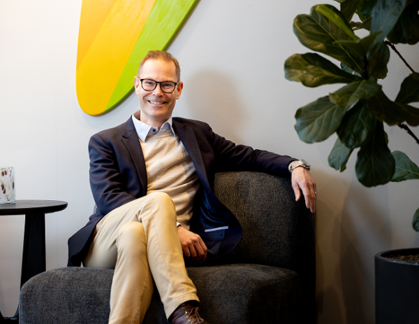 En man sitter i en fåtölj i en kontorsmiljö och ler mot kameran.
