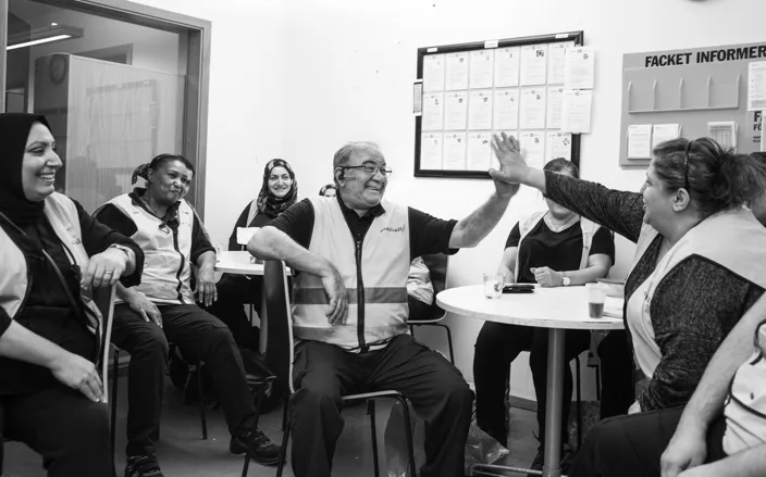 En grupp arbetskamrater sitter runt ett bord i ett personalrum. En man och en kvinna ger varandra en high five medan de andra runt bordet ler och ser på. På väggen bakom dem sitter en anslagstavla med information och dokument.