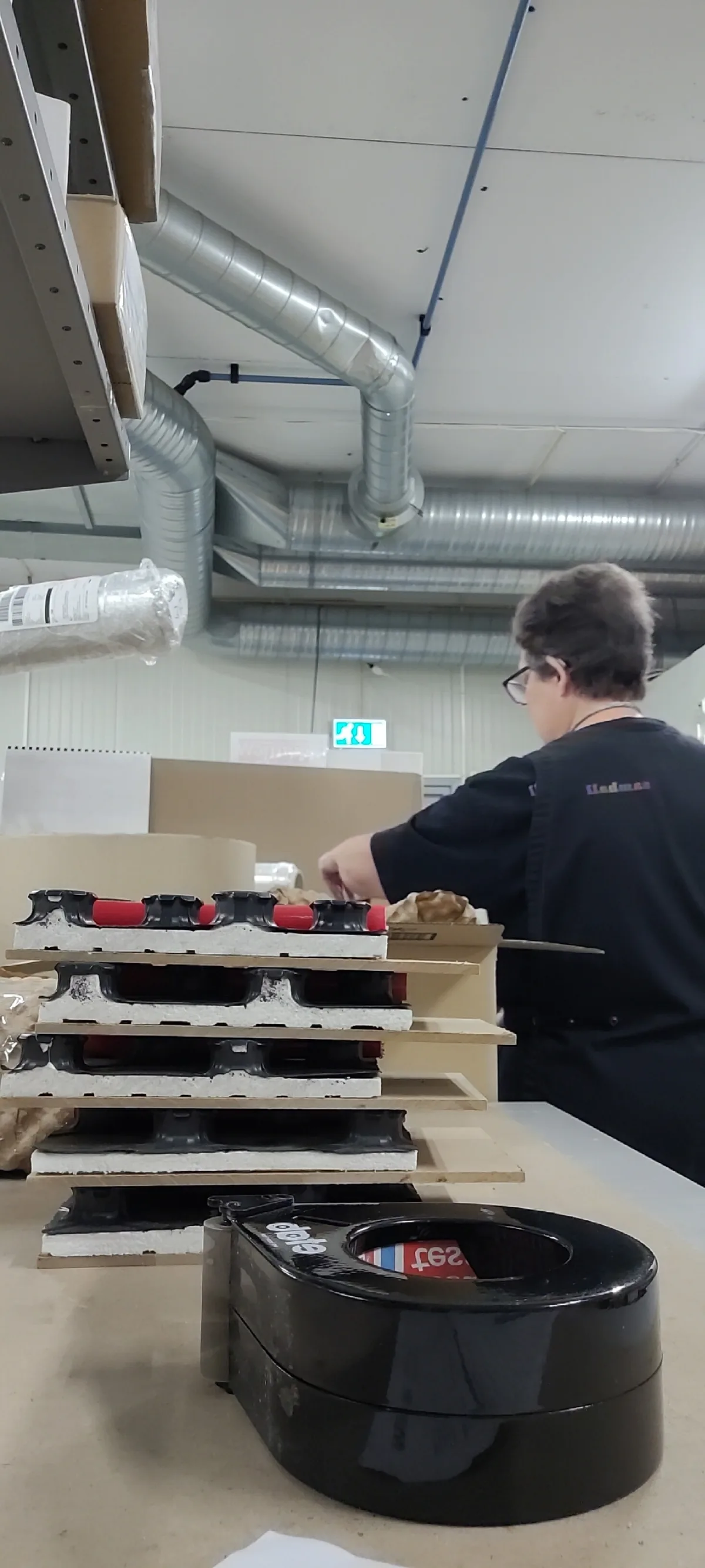 Person står vid ett arbetsbord i en industriell arbetslokal och packar föremål. I förgrunden syns staplade förpackningsinsatser och en svart tejphållare på bordet. I bakgrunden finns ventilationsrör i taket och en nödutgångsskylt.