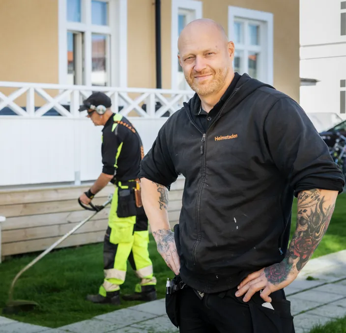 Niklas Eklund, fastighetsskötare på Heimstaden, poserar framför kamran samtsiigt som en medarbetare klipper gräset i bakgrunden.