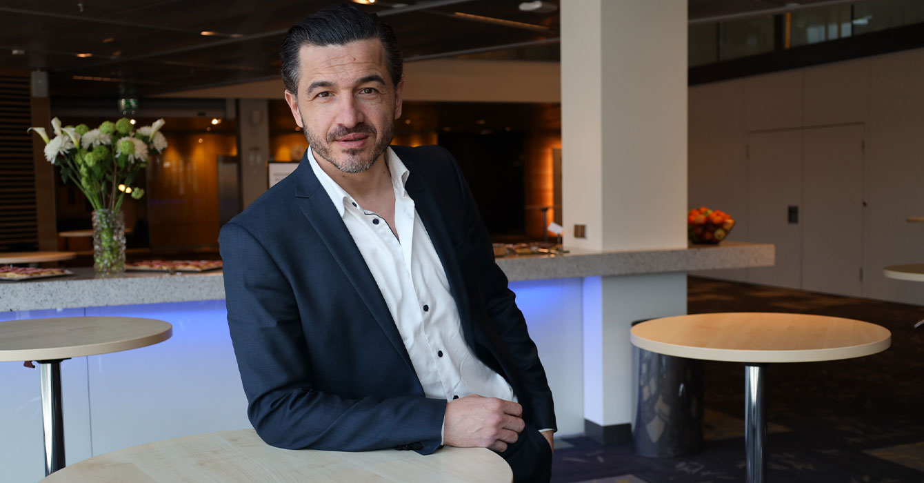 En man i kostym och svart hår står lutad mot ett småbord och tittar in i kameran.