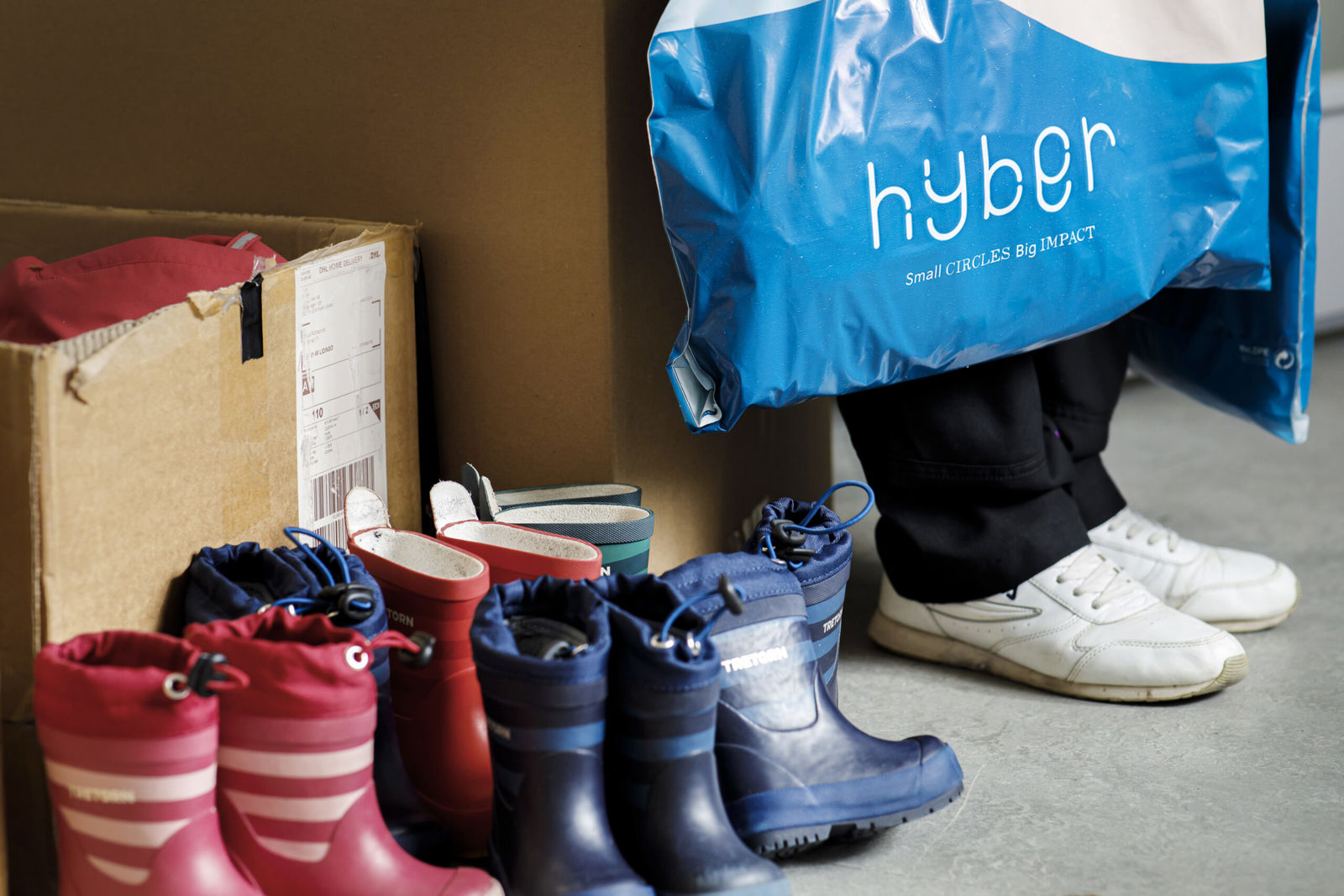 Flera par färgglada barnstövlar står uppradade på golvet bredvid en kartong. En person med vita sneakers håller en blå påse.