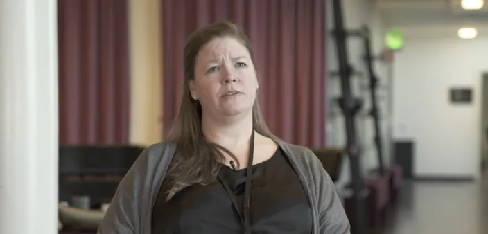 Veronica Jakobsson står inomhus och talar, klädd i svart topp och grå kofta med en namnbricka runt halsen. I bakgrunden syns röda gardiner och en ljus, öppen miljö.