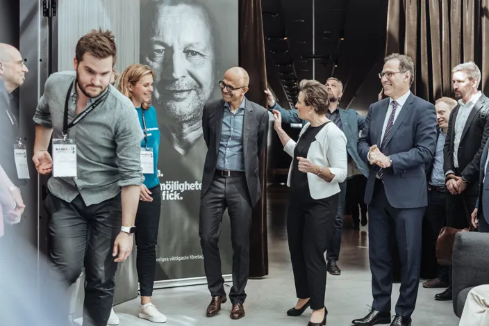 En grupp personer med namnskyltar står i en konferensmiljö och samtalar. I bakgrunden syns en stor affisch med ett porträtt, och några deltagare rör sig genom en korridor.