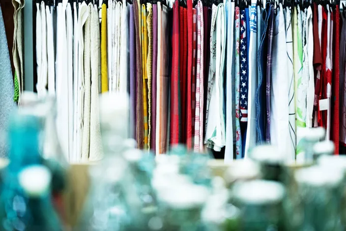 En rad färgglada tygstycken hänger tätt bredvid varandra på en stång. I förgrunden syns suddiga glasburkar eller flaskor, medan tygerna i bakgrunden visar olika färger och mönster.