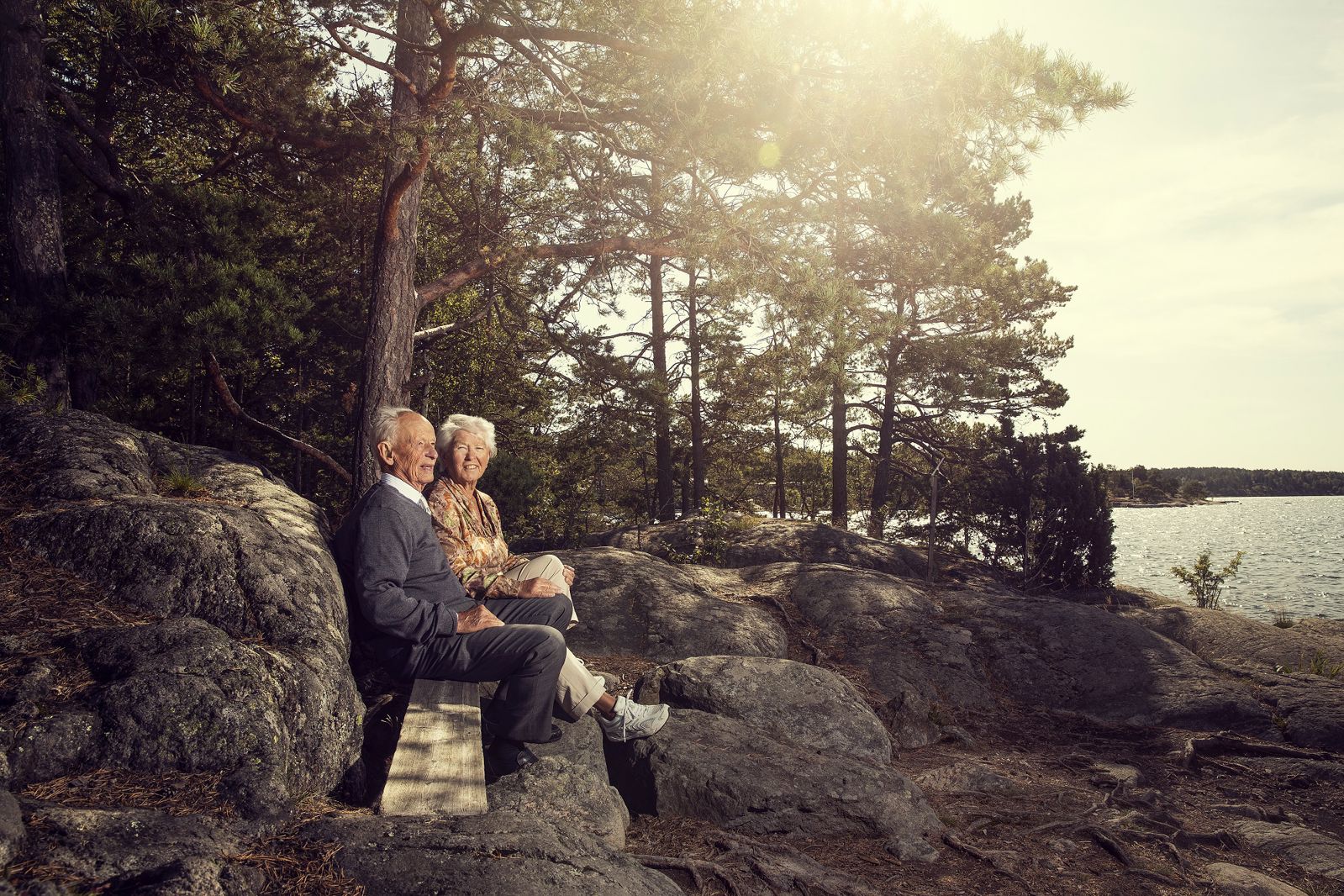 Lillemor och Björn sitter tillsammans på en träbänk bland klippor och tallar vid vattnet i varmt kvällsljus. De tittar ut över havet medan solen lyser genom träden bakom dem.