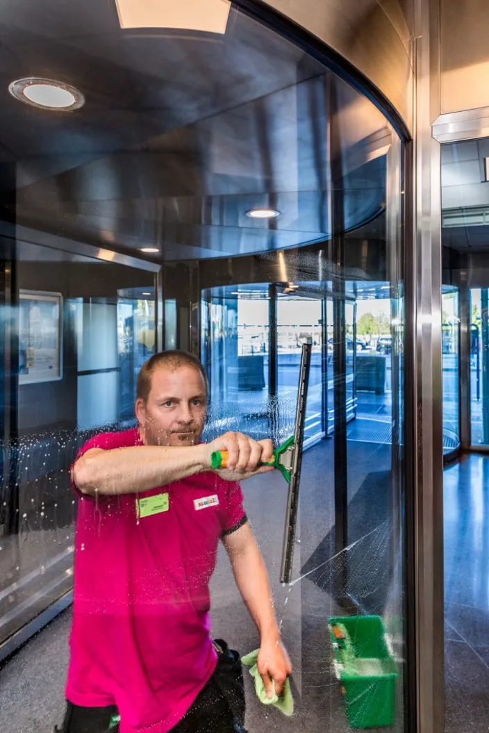 En man i rosa arbetskläder putsar en glasdörr med en fönsterskrapa i en entré med snurrdörr och glaspartier.