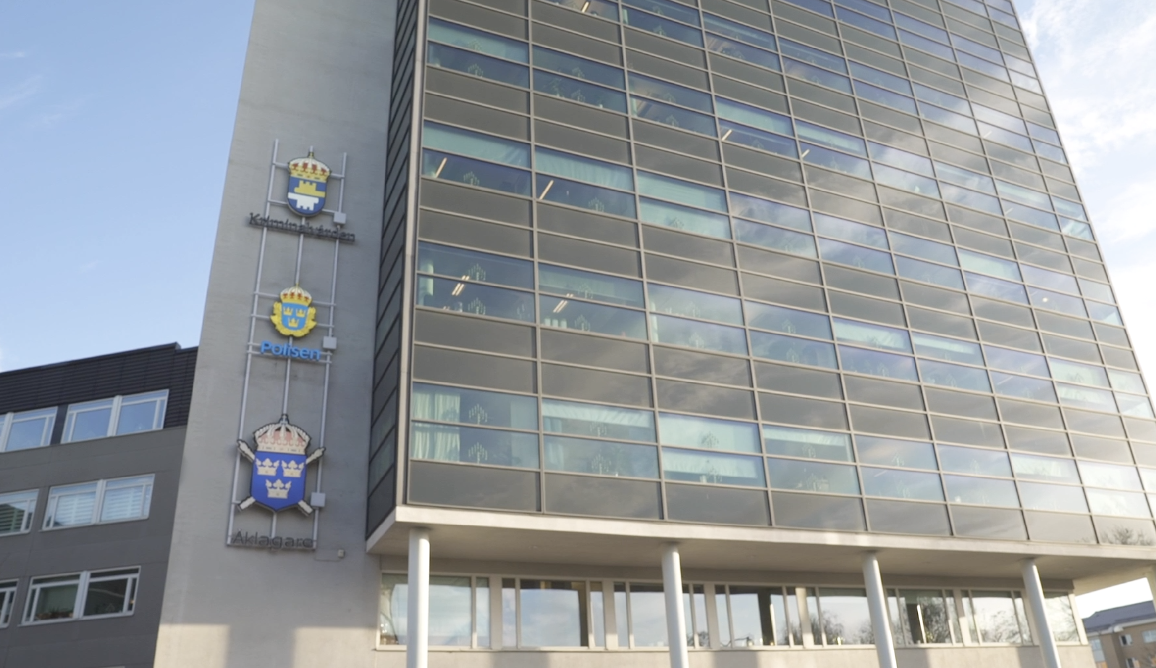 En modern kontorsbyggnad med glasfasad sedd underifrån. På byggnadens sida sitter tre vapensköldar och skyltar med texten ”Polisen”, ”Åklagare” och "kriminalvården", vilket visar att huset rymmer polis, åklagarmyndighet samt kriminalvården.