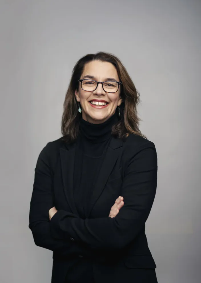 Porträtt av Sara Revell Ford framför en grå bakgrund. Hon bär svart kavaj och har armarna i kors. 