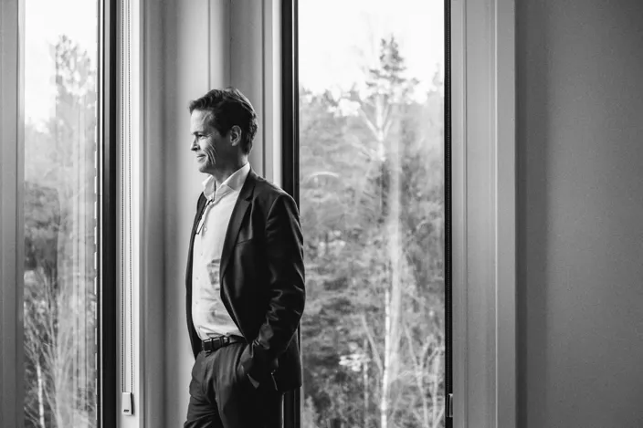 En svartvit bild av en man i kostym som står vid ett stort fönster med händerna i fickorna och blickar ut mot träd utanför.