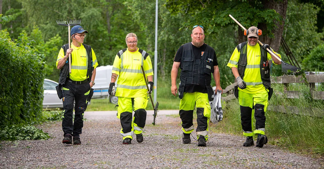 Samhalls medarbetare på fastighetsavdelningen i Strängnäs går på en grusväg mot kameran, De bär på redskap och har arbetskläder i reflex.