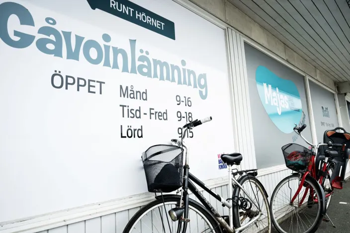 Skyltfönster med texten ”Gåvoinlämning” och öppettider (måndag 9–16, tisdag–fredag 9–18, lördag 9–15). Framför väggen står två cyklar parkerade, och i fönstret syns även logotypen ”Majas”.