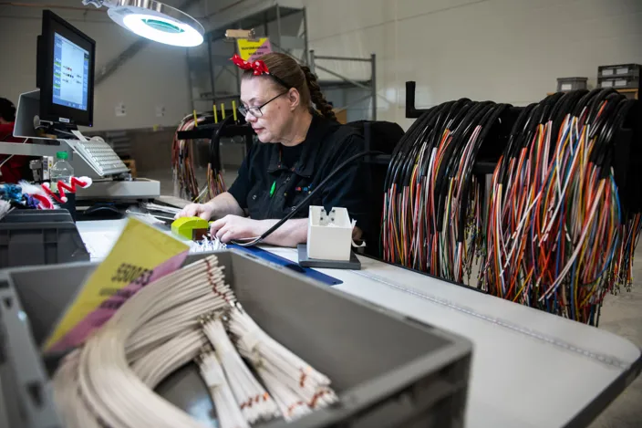 Carina sitter vid ett skrivbord och arbetar. Bredvid henne hänger massor av sladdar. 