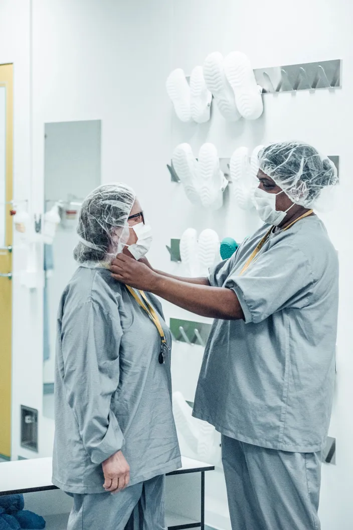 Två personer i skyddskläder, munskydd och hårskydd står i en steril miljö där den ena justerar den andras skyddsutrustning. I bakgrunden hänger vita arbetsskor på väggen.