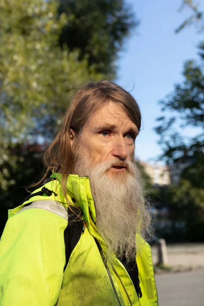 En man med långt hår och långt vitt skägg står utomhus och tittar åt sidan. Han bär en neonfärgad jacka och har träd och blå himmel i bakgrunden.
