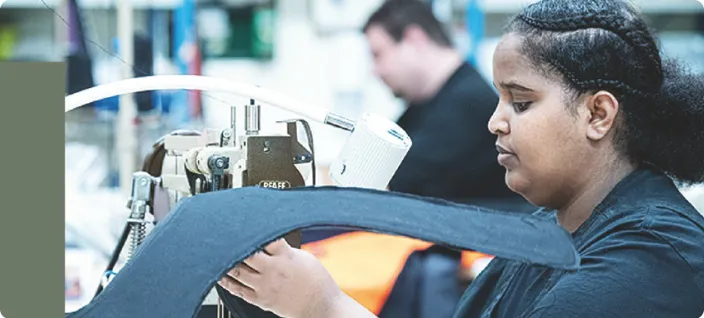 Samhallsmedarbetare arbetar vid en symaskin och syr på ett kraftigt tyg i en produktionsmiljö.