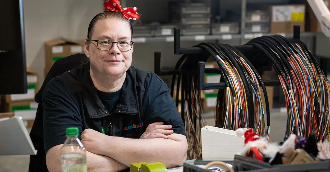 Foto av person som sitter lutad vid ett arbetsbord, lutandes med armarna i kors. Personer bär en röd rosett på huvudet och glasögon.