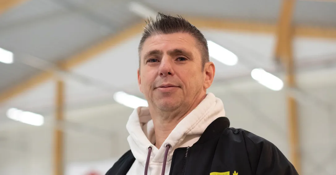 Porträtt av en mans ansikte som tittar in i kameran. Har kort grått/svart hår och svart tröja.