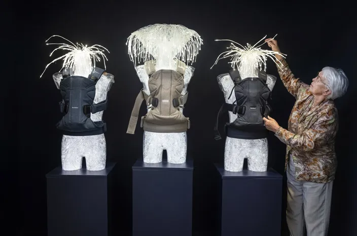 Tre dockliknande överkroppar står på piedestaler mot en svart bakgrund och bär olika modeller av bärselar. Lillemor står bredvid och justerar den högra bärselen med handen, medan de vita, trådliknande “huvudena” på figurerna spretar uppåt som hår.