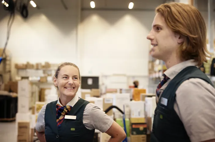 Bild på två personer som pratar med varandra inne på Systembolagets lager. En kvinna och en långhårig man.