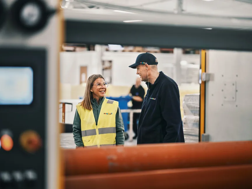 En kvinna i gul varselväst och en man i svart keps och svart jacka samtalar och ler i en industrihall.