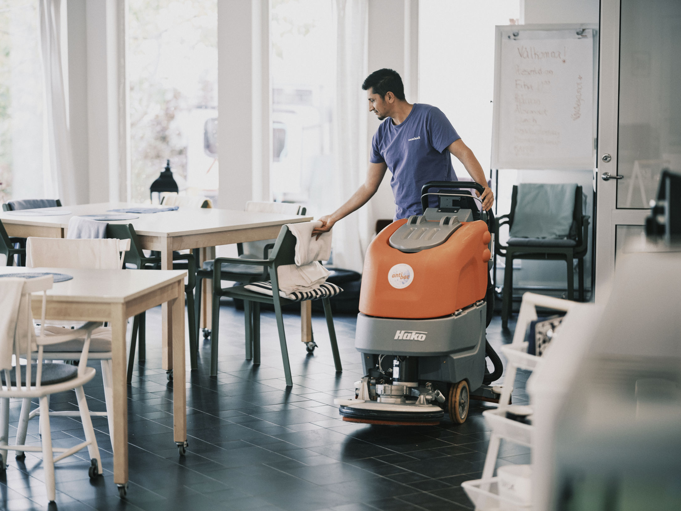 Äldreboende i Sköndal. Medarbetare Jama rättar till en stol i matsalen och hanterar en stor skurmaskin. Jama är mörkhårig och har lila Samhall-t-shirt.