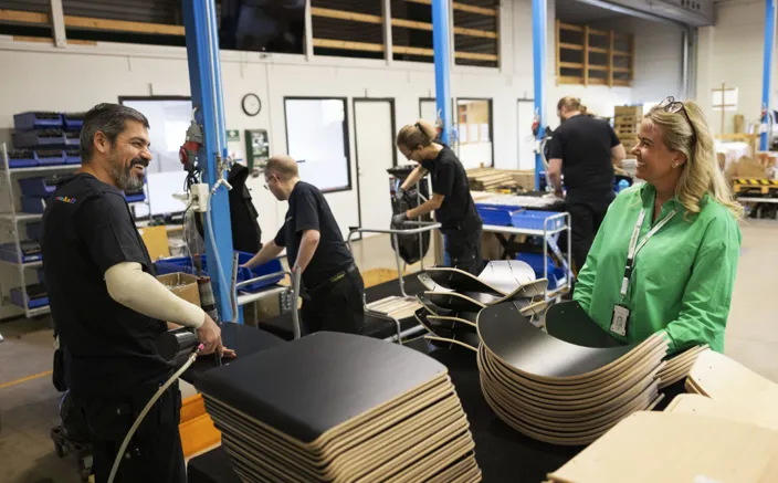 Samhallmedarbetare med svarta arbetskläder arbetar med material i verkstad. Carlotte klädd i grönt ler åt en medarbetare.