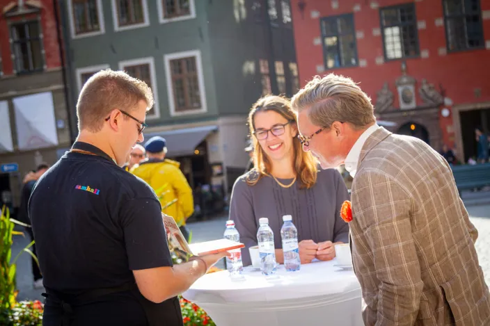 Tre personer står runt ett högt bord utomhus i solsken och samtalar. På bordet finns vattenflaskor och kaffekoppar, och i bakgrunden syns färgglada byggnader på ett torg.