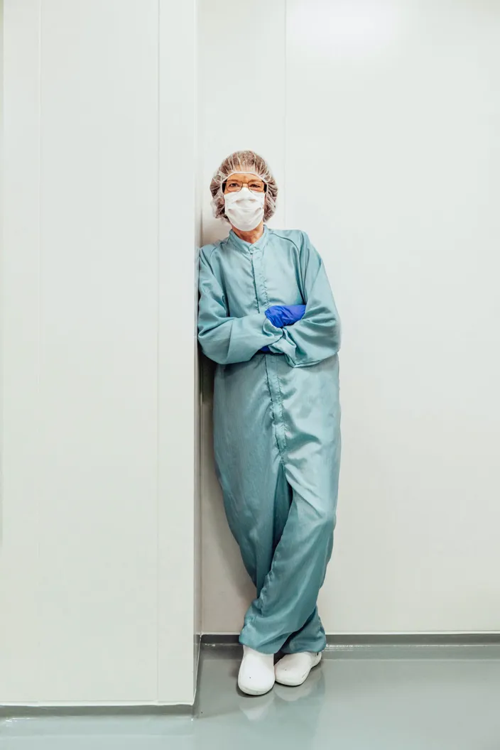 En person i skyddsdräkt, munskydd, hårskydd och blå handskar står lutad mot en vit vägg i en steril miljö, med armarna korsade.