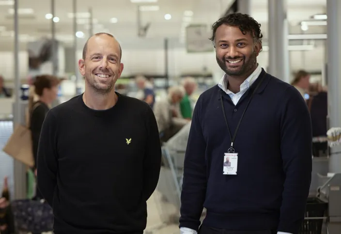 Två män står i en Sytembolaget-butik och tittar in i kameran. Båda ler.