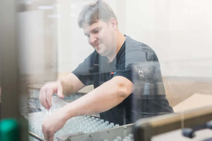 Andreas Gräntz matar flaskor i en maskin. Fotot är taget genom en glasruta.