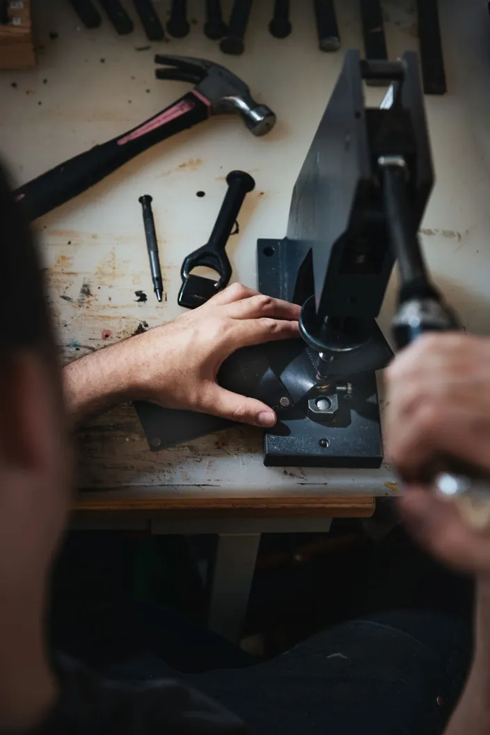En person arbetar vid en arbetsbänk och använder en handdriven press för att bearbeta en liten metalldel. På bordet ligger verktyg som hammare, skruvmejsel och andra metallverktyg utspridda.
