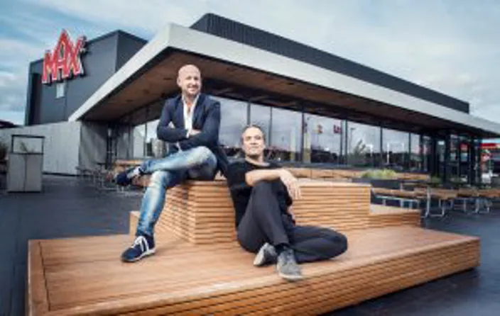 Christoffer Bergfors och Kaj Török sitter på en träbänk utanför en MAX-restaurang med den röda MAX-logotypen synlig på byggnaden bakom dem.