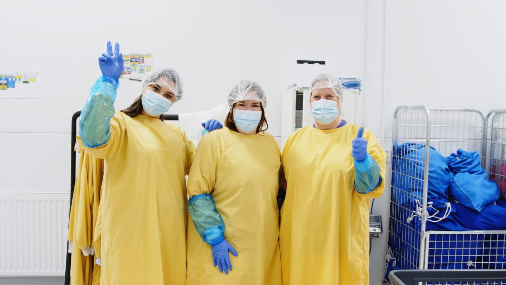 Tre samhallsmedarbetare i skyddskläder, hårnät och munskydd står i en hygienisk arbetsmiljö och gör tummen upp mot kameran.