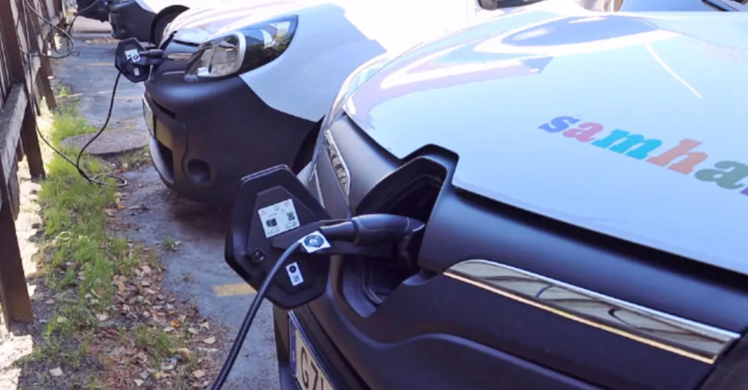 Närbild på en vit elbil som står och laddar med en sladd inkopplad. Man ser Samhall-loggan på motorhuven.