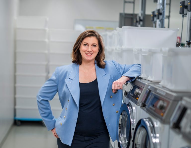 Porträttbild på Jessica Bederoff Stenvad, Hållbarhetschef, Samhall, Stockholm. Fotograferad på ett tvätteri/i en tvättstuga. Jessica har brunt hår, mörkblått linne och en ljusblå kavaj.