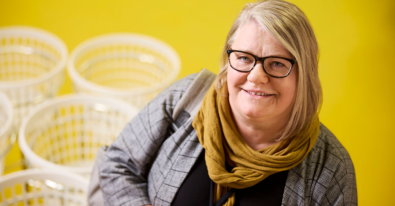 Porträtt på Agneta som har grått hår, svarta glasögon och tittar in i kameran leendes. Bakgrunden är gul och har några vita tvättkorgar.