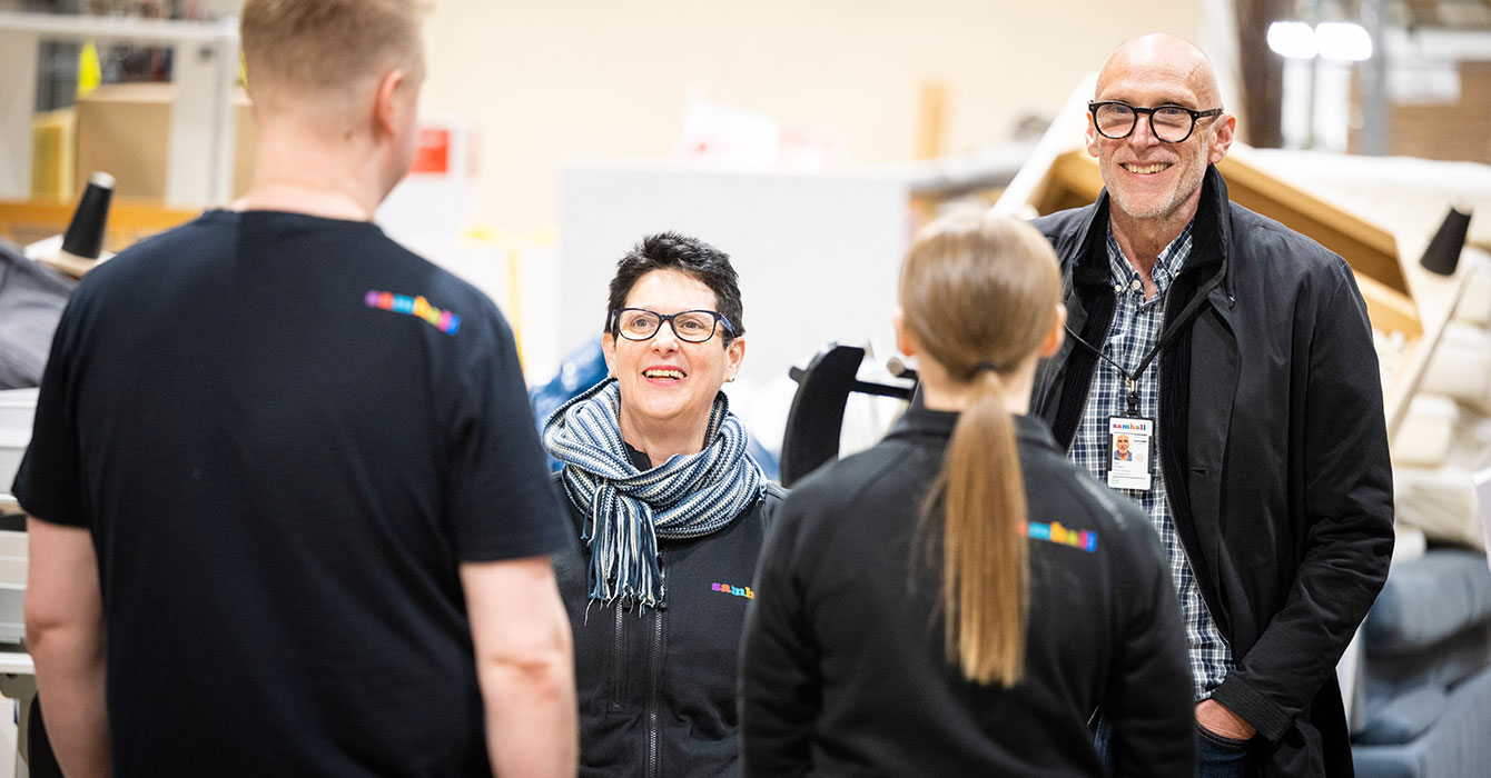 En kvinna och en man står vänd mot två Samhallmedarbetare och pratar, Man ser ryggen på medarbetarna. Kvinnan och manen ler och bär glasögon.