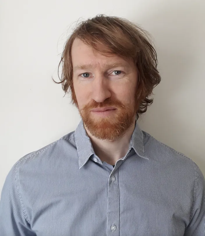 Johan Stenmark tittar in i kameran och bär en blå skjorta. Bakgrunden är vit och hans hår och skägg är brunt.