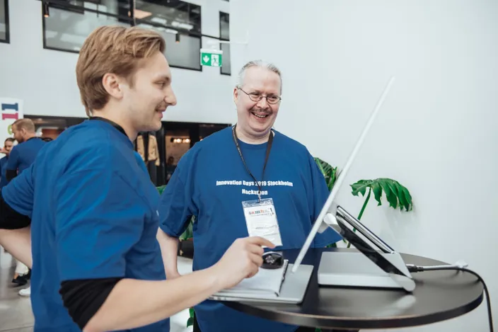Två deltagare i blå t-shirts och namnskyltar står vid ett bord och pratar, medan de tittar på en dator i en ljus konferensmiljö.