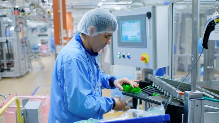 En person i blå skyddsrock och hårnät arbetar vid en maskin i en industriell produktionsmiljö. Personen placerar gröna plastdetaljer på ett transportband medan en kontrollpanel med display syns bredvid.