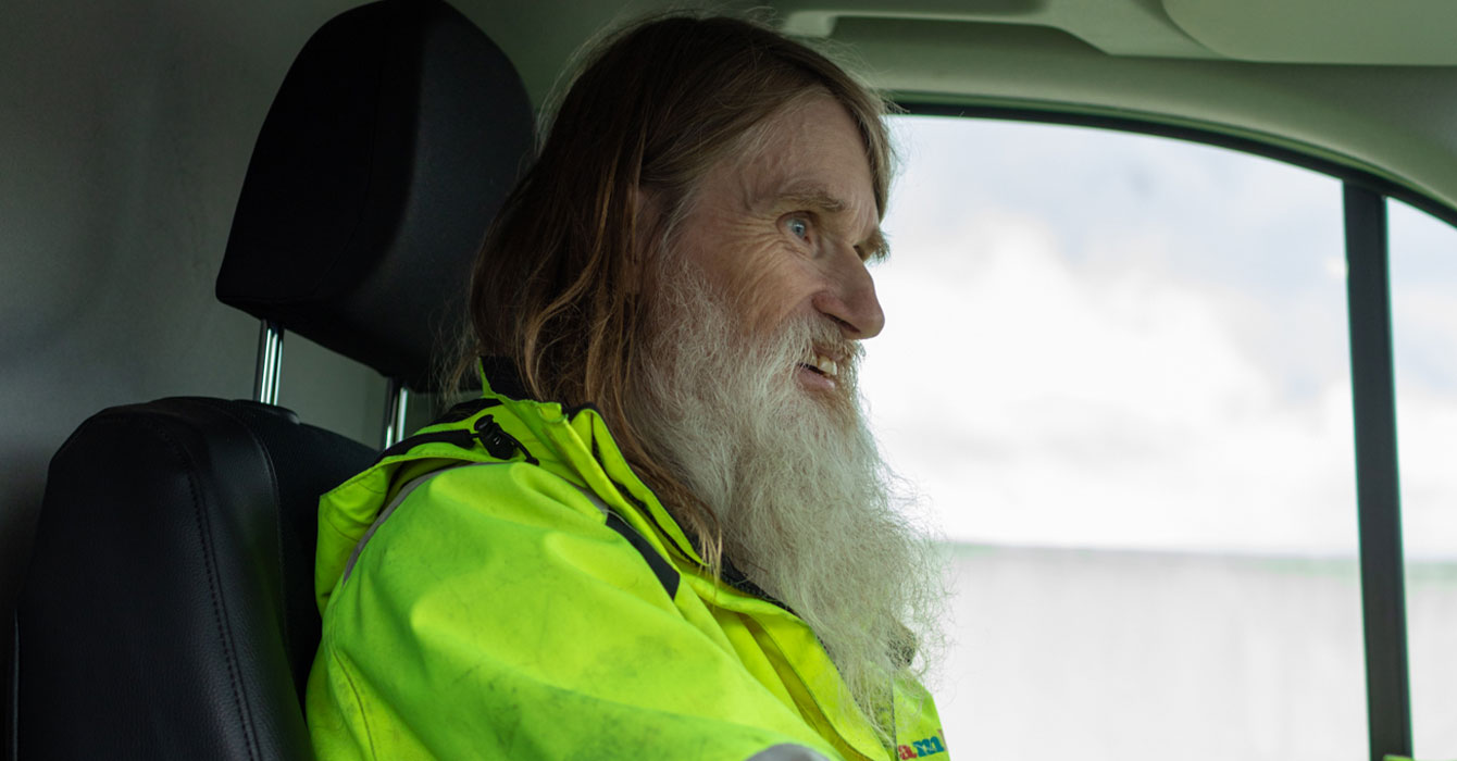 Bild på en man i signalfärgad jacka som kör bil. Mannen har grått långt hår.