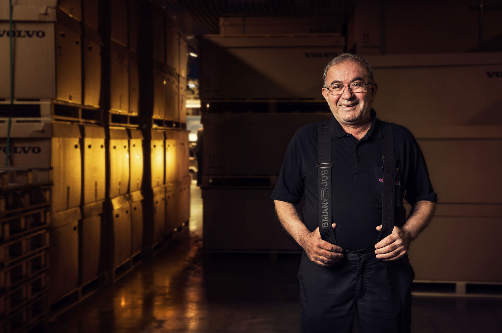 En leende man står i en lagerlokal mellan höga staplar av stora trälådor märkta ”Volvo”. Han bär mörka arbetskläder med hängslen och är upplyst av varmt ljus i den annars mörka industrimiljön.