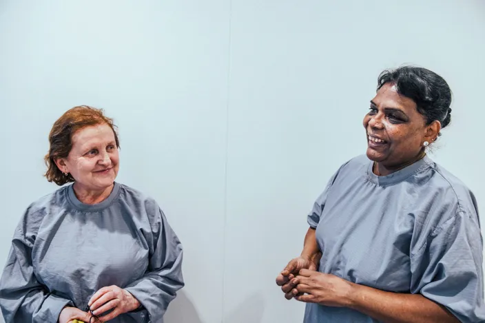 Två personer i arbetskläder står mot en ljus vägg och ler mot varandra under ett samtal i en ren och avskalad miljö.