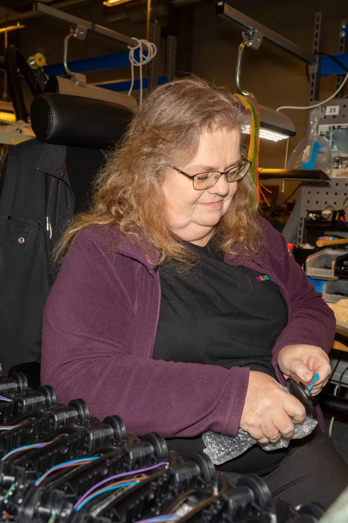 En kvinna med glasögon sitter vid en arbetsstation i en verkstadsmiljö och monterar eller hanterar en mindre komponent. Hon bär en mörk tröja och lila kofta, och framför henne ligger flera svarta delar uppradade på ett bord. I bakgrunden syns arbetsbelysning och hyllor med utrustning.