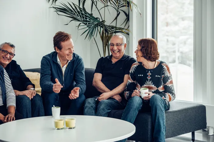 Fyra personer sitter tillsammans i en soffa i en ljus lounge och skrattar under ett samtal. På bordet framför dem står kaffekoppar och ljuslyktor, och en stor växt syns i bakgrunden.