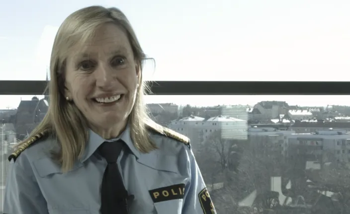 Carin Götblad i polisuniform med slips och axelklaffar står framför ett stort fönster med utsikt över en stadsmiljö. På uniformen syns texten ”POLIS”.