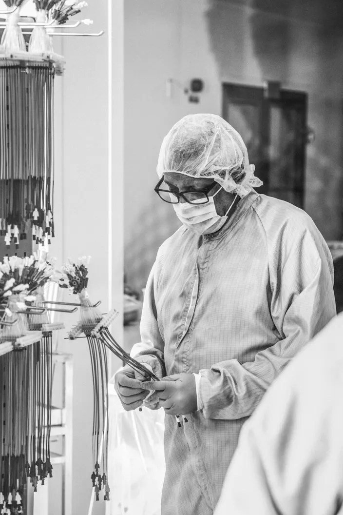 En svartvit bild av en person i skyddskläder, hårnät, munskydd och handskar som står i en produktionsmiljö och inspekterar buntar av slangar.