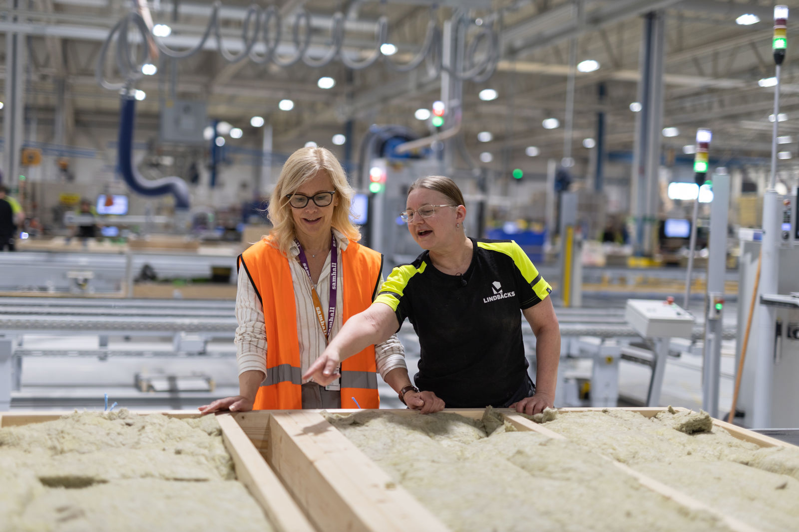 Elina, områdeschef, och Helen, byggarbetare. De jobbar vid ett bord med träreglar som har isolering mellan. Helen pekar mot bordet. Elina har varselväst, Helen har Lindbäck-kläder..