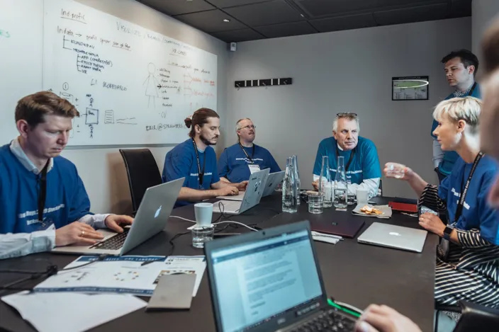 Ett möte pågår i ett konferensrum där flera personer i blå t-shirts sitter runt ett bord med laptops och anteckningar. På väggen bakom syns en whiteboard med skisser och text.